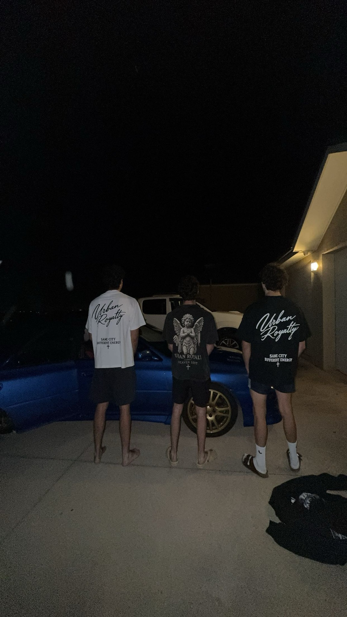 Three people wearing the Urban Royalty T-shirts beside the blue car at night